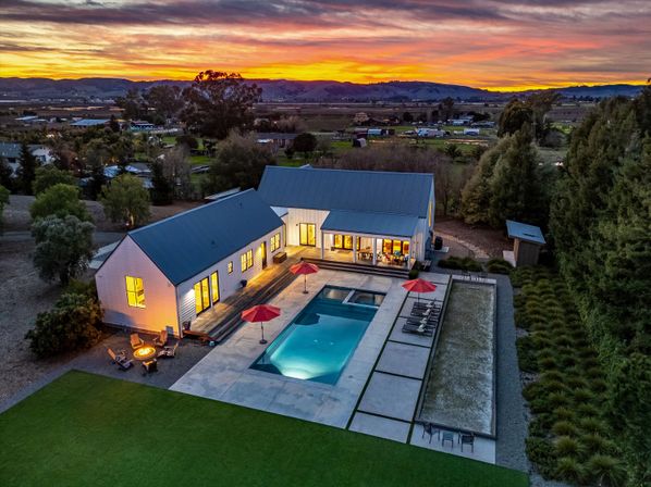 Aerial dusk view of a modern farmhouse in the countryside with glowing windows, a concrete patio and rectangular swimming pool with red umbrellas, lounge chairs and a fire pit, surrounded by fields and rolling hills at sunset.