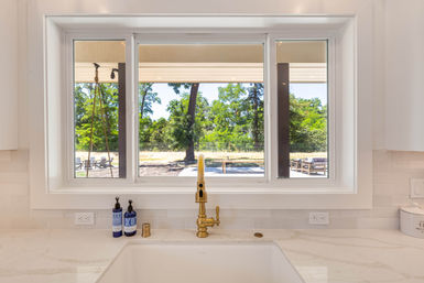 Sunlit modern kitchen sink with a brass faucet and marble countertop, blue soap bottles on the sill, looking through a large window onto a sunny backyard patio and trees.