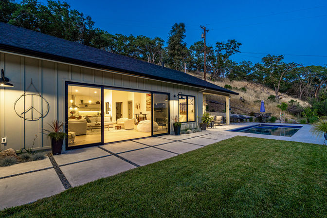 Modern single-story hillside home at dusk with warm open-plan living visible through large sliding glass doors, concrete paver backyard patio, manicured lawn, rectangular swimming pool and outdoor seating.