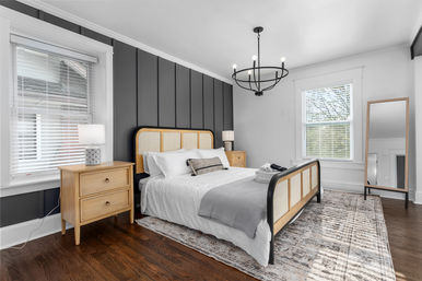 Sunlit modern bedroom interior with gray paneled accent wall, rattan-framed bed, wooden nightstands, black chandelier, area rug, and hardwood floors.