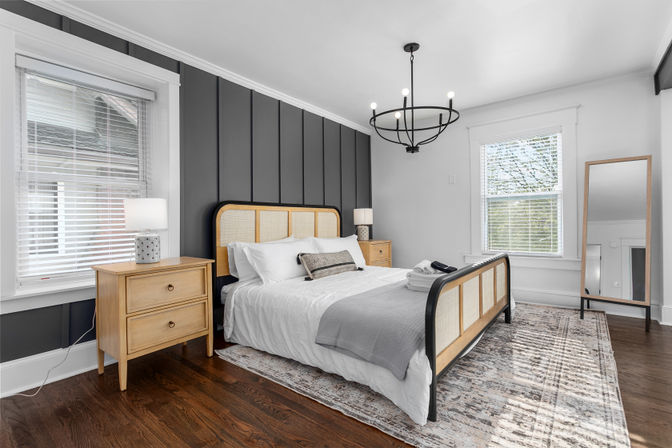 Sunlit modern bedroom interior with gray paneled accent wall, rattan-framed bed, wooden nightstands, black chandelier, area rug, and hardwood floors.