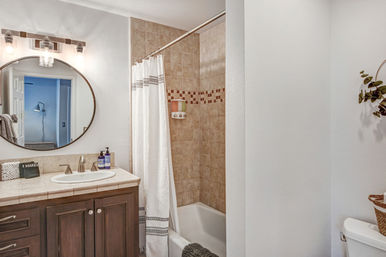 Cozy residential bathroom with dark wood vanity, round mirror and sconce lights, tiled bathtub/shower with mosaic border, white patterned shower curtain and chrome fixtures