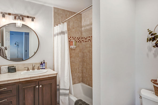 Cozy residential bathroom with dark wood vanity, round mirror and sconce lights, tiled bathtub/shower with mosaic border, white patterned shower curtain and chrome fixtures