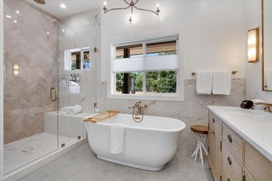 Bright modern bathroom with white freestanding tub and wooden caddy, glass-enclosed shower with herringbone marble tile, light wood vanity, gold fixtures and a window view of trees.