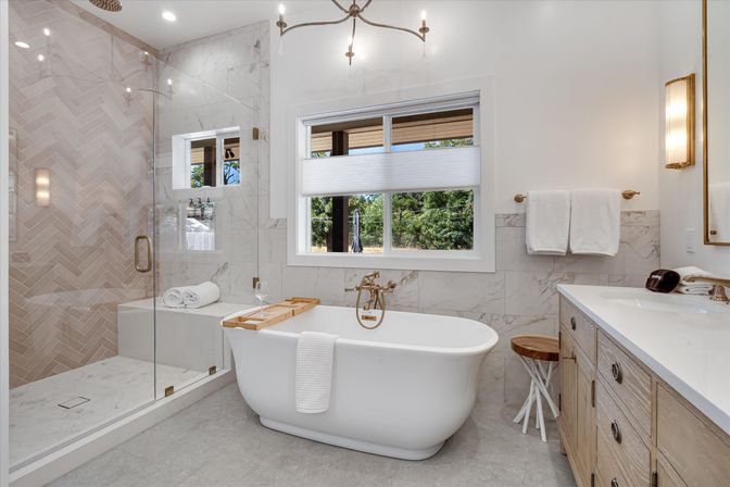 Bright modern bathroom with white freestanding tub and wooden caddy, glass-enclosed shower with herringbone marble tile, light wood vanity, gold fixtures and a window view of trees.