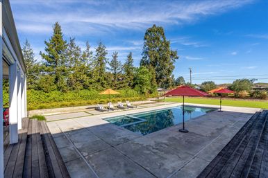Sunny modern residential backyard with rectangular outdoor swimming pool, concrete patio, red and orange umbrellas, lounge chairs, green lawn and tall trees under a clear blue sky