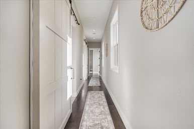 Bright narrow residential hallway interior with dark hardwood floors, light gray walls, two long patterned runner rugs, white sliding barn door on the left, window and woven wall decor on the right, and recessed ceiling lights.