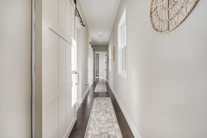 Bright narrow residential hallway interior with dark hardwood floors, light gray walls, two long patterned runner rugs, white sliding barn door on the left, window and woven wall decor on the right, and recessed ceiling lights.