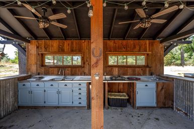 Rustic outdoor kitchen in a covered pavilion with blue lower cabinets, stainless counters, double sinks, corrugated metal panels, ceiling fans, string lights, and a horseshoe-decorated wooden post.
