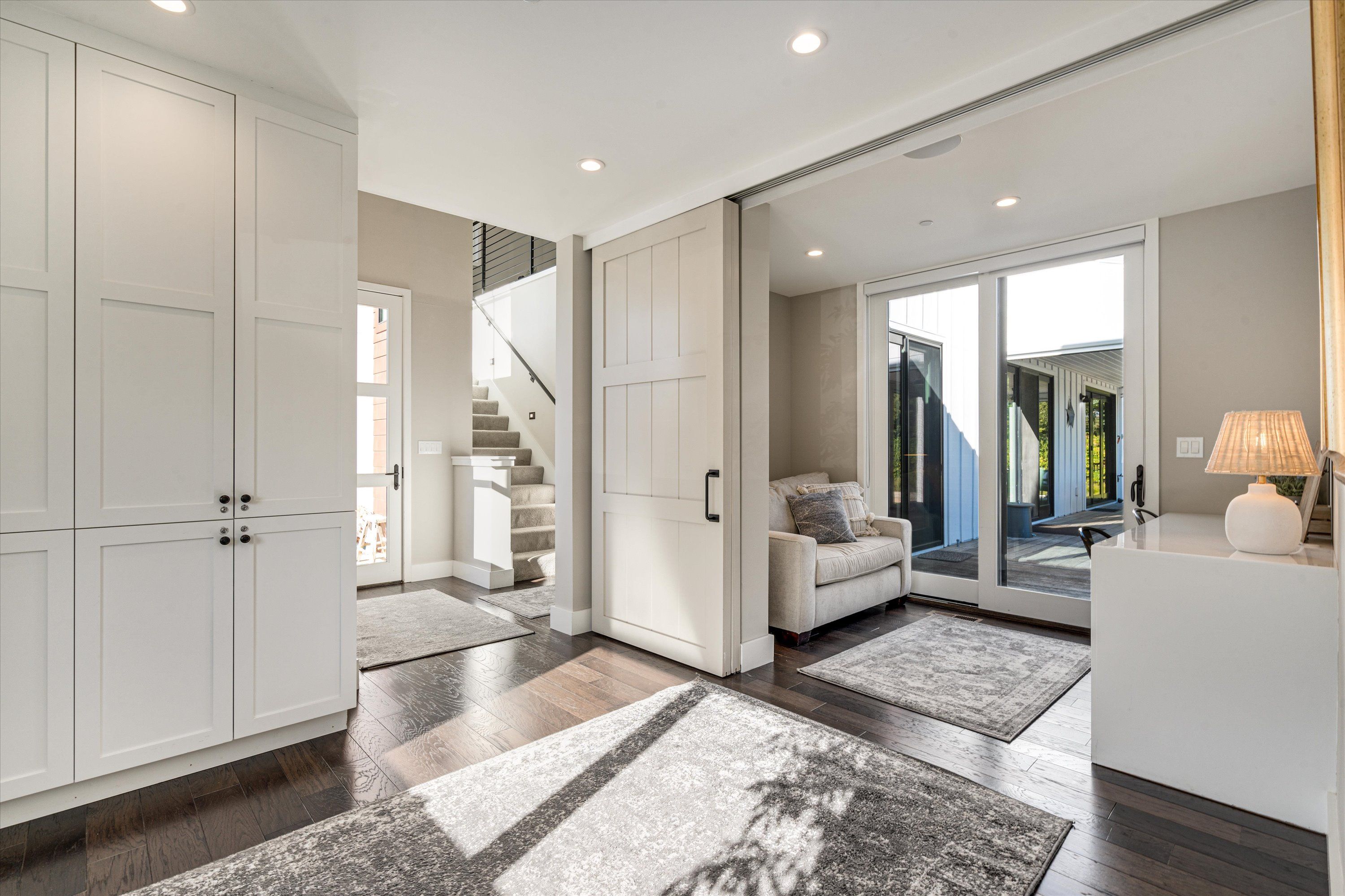 Sunlit modern foyer with white built-in cabinets, dark hardwood floors, sliding barn door, staircase, cozy sitting nook and glass patio doors.