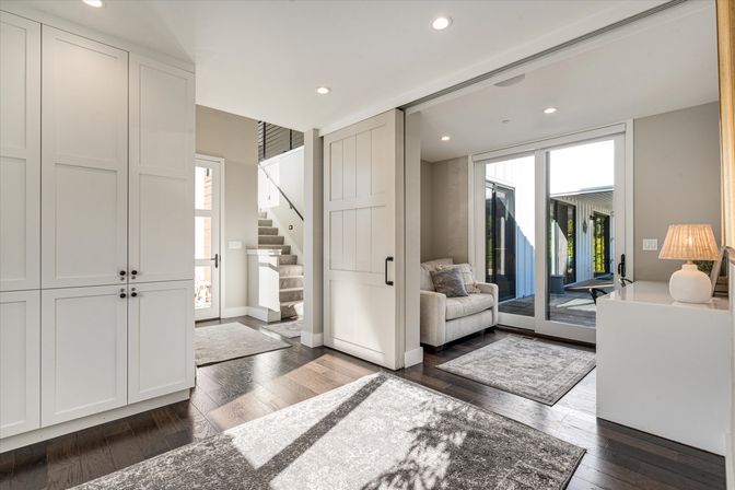 Sunlit modern foyer with white built-in cabinets, dark hardwood floors, sliding barn door, staircase, cozy sitting nook and glass patio doors.
