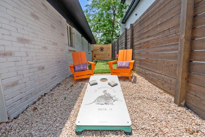 Narrow sunny Austin backyard patio with a cornhole board labeled 'Austin, TX', two bright orange Adirondack chairs with 'relax' pillows on crushed gravel between a white brick house and a wooden privacy fence.