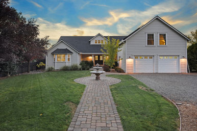 Inviting suburban gray two-story house at dusk with a brick walkway leading to a central stone fountain, manicured green lawn, lit windows and a three-car garage