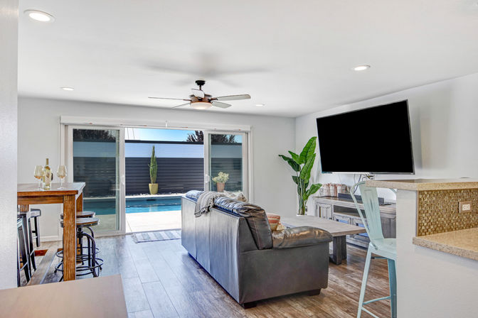 Sunlit open-plan living room with dark leather sofa facing a wall-mounted TV, wood-look floors, breakfast bar with metal stools and sliding glass doors opening to a backyard pool and potted plants.