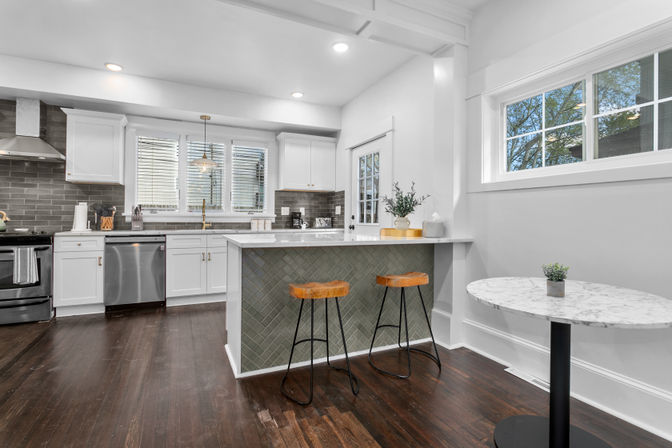 Bright modern open-concept kitchen with white cabinets, gray subway-tile backsplash, stainless steel appliances, marble-topped breakfast bar with two wooden stools, small round marble cafe table, and dark hardwood floors.