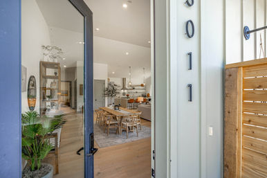 Open glass front door revealing a bright, modern open-plan home interior with a wooden dining table and chairs on light oak floors, potted plants, pendant-lit kitchen and cozy living area.