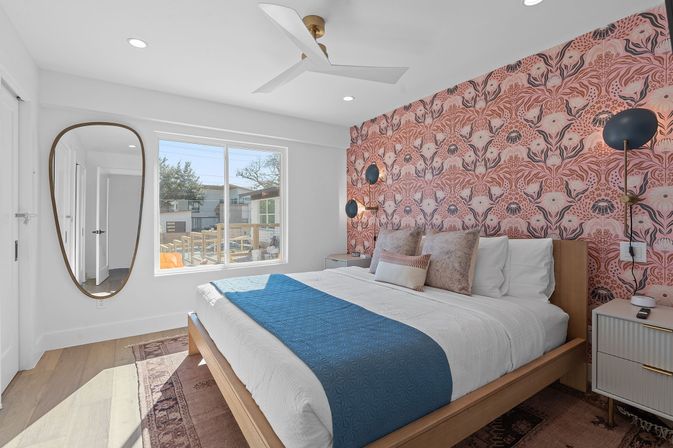 Sunlit modern bedroom with pink floral accent wall, wooden platform bed with blue runner, oval wall mirror, brass sconces, ceiling fan, and large window overlooking a residential street.