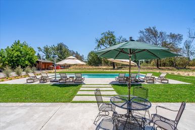 Spacious residential backyard pool with lounge chairs and large green umbrellas on a concrete patio, metal bistro table in the foreground, manicured lawn and trees under a clear blue sky
