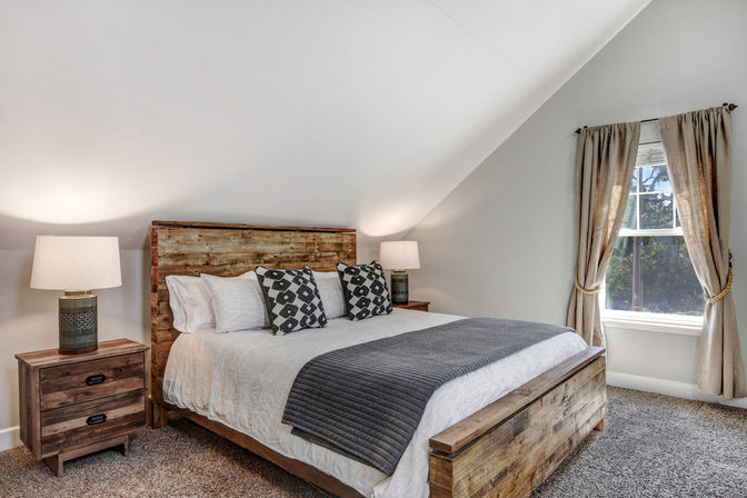 Cozy rustic loft bedroom with sloped ceiling, reclaimed-wood bedframe, neutral bedding, gray quilt and patterned pillows, two nightstands with lamps, carpeted floor and a curtained window with natural light.