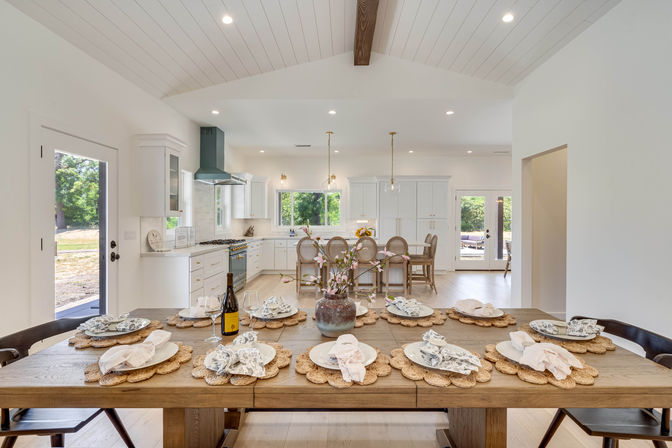 Bright open-concept farmhouse kitchen and dining area with vaulted shiplap ceiling, wooden table set for eight with woven placemats and floral napkins, white cabinets, island with rattan stools, pendant lights, and large windows overlooking green yard.