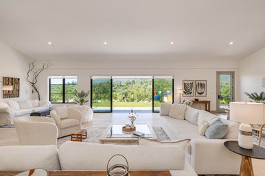 Bright modern open-plan living room with cream sofas and armchairs, glass coffee table and panoramic sliding doors opening to a lush backyard and hillside view.