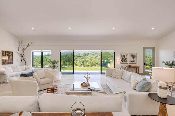 Bright modern open-plan living room with cream sofas and armchairs, glass coffee table and panoramic sliding doors opening to a lush backyard and hillside view.