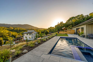 Sunset-lit backyard with a rectangular pool and concrete patio beside a modern hillside home, overlooking green rolling hills and oak-studded landscape.