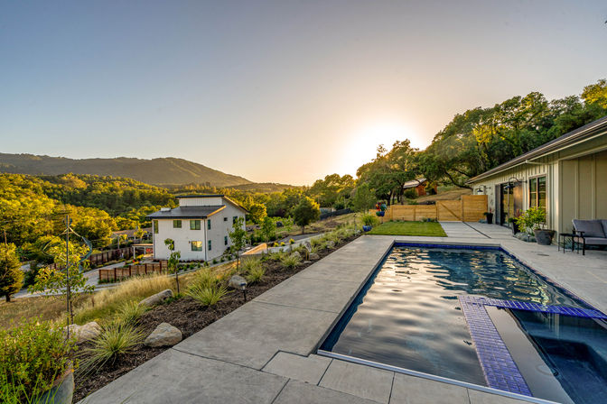 Sunset-lit backyard with a rectangular pool and concrete patio beside a modern hillside home, overlooking green rolling hills and oak-studded landscape.