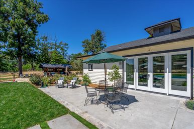 Sunlit residential backyard patio with metal dining set and green umbrella on a concrete terrace, lounge chairs, manicured lawn, private pool visible through glass patio doors, surrounding trees and small gazebo under a clear blue sky.