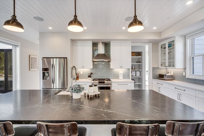 Bright modern kitchen with a large dark marble island and bar stools, white shaker cabinets, stainless steel refrigerator and range, gray subway-tile backsplash, pendant lights and open shelving