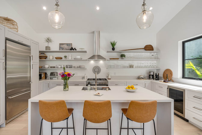 Bright modern white kitchen with a large island, three tan leather bar stools, stainless refrigerator and range hood, open floating shelves, glass pendant lights, vase of colorful flowers and a bowl of lemons on the counter.