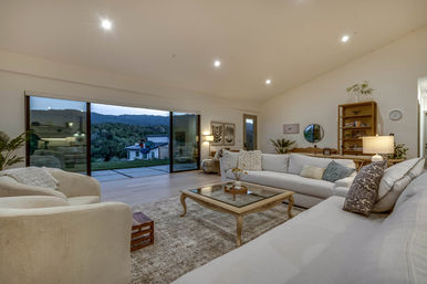 Cozy open-plan living room with neutral sofas and armchairs around a glass-top wooden coffee table, bookshelf and warm lamps, opening through floor-to-ceiling sliding glass doors to a patio and dusk mountain view.