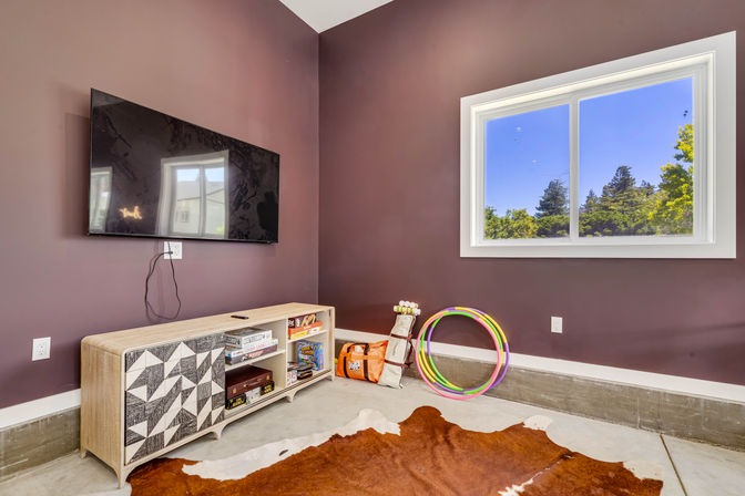 Modern playroom corner with wall-mounted flat-screen TV on plum walls, light wood storage cabinet with board games, colorful hula hoops and toy bag on concrete floor, cowhide rug and bright window view of trees