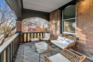 Cozy covered brick porch with wicker seating, cushioned bench and chairs, glass coffee table on a striped rug, wooden railing and autumn trees beyond