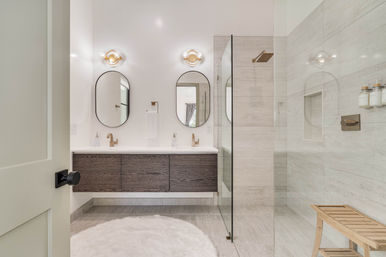 Spa-like modern bathroom with floating dark wood double vanity, two oval mirrors, brass faucets, walk-in glass shower with beige tile, wooden bench and round white shag rug