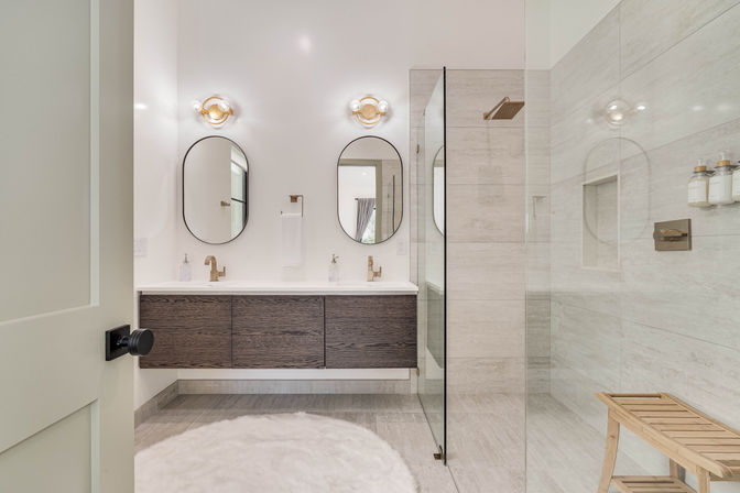 Spa-like modern bathroom with floating dark wood double vanity, two oval mirrors, brass faucets, walk-in glass shower with beige tile, wooden bench and round white shag rug