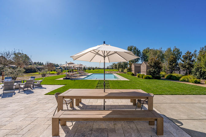 Sunny modern backyard patio with wooden picnic table and umbrella overlooking a rectangular pool, lounge chairs, and manicured lawn
