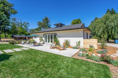 Sunlit backyard patio of a modern white cottage with concrete terrace, wicker lounge chairs with striped cushions, dining table under a green umbrella, manicured lawn, flower beds, wooden privacy fence and mature trees under a clear blue sky