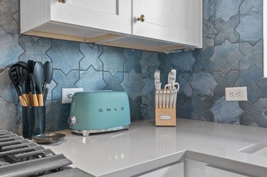 Bright modern kitchen countertop with teal retro toaster, wooden knife block, black cooking utensils in a jar, white cabinets, and blue geometric tile backsplash.