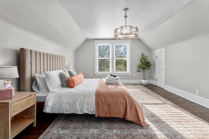 Sunlit cozy attic bedroom with sloped ceiling, tufted beige headboard, crisp white bedding, coral throw and lumbar pillow, wooden nightstand and lamp, modern brass chandelier, potted plant by the window, and dark hardwood floors over a patterned rug.