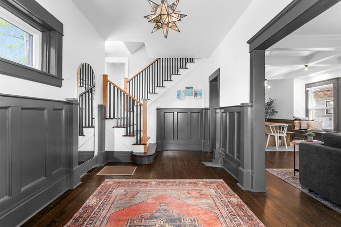 Inviting craftsman-style home entryway with dark gray wainscoting, hardwood floors, patterned red area rug, wooden staircase with black spindles and a star pendant light opening to a bright living and dining area.