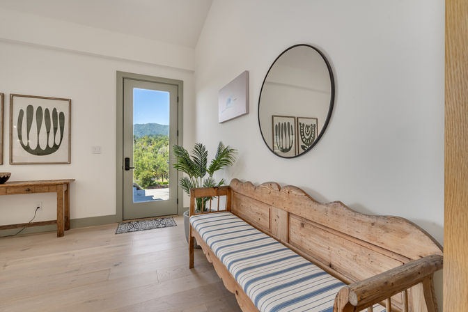 Sunlit foyer with rustic wooden bench topped by a blue-striped cushion, round wall mirror, potted palms, and a glass door framing green hills and mountain views