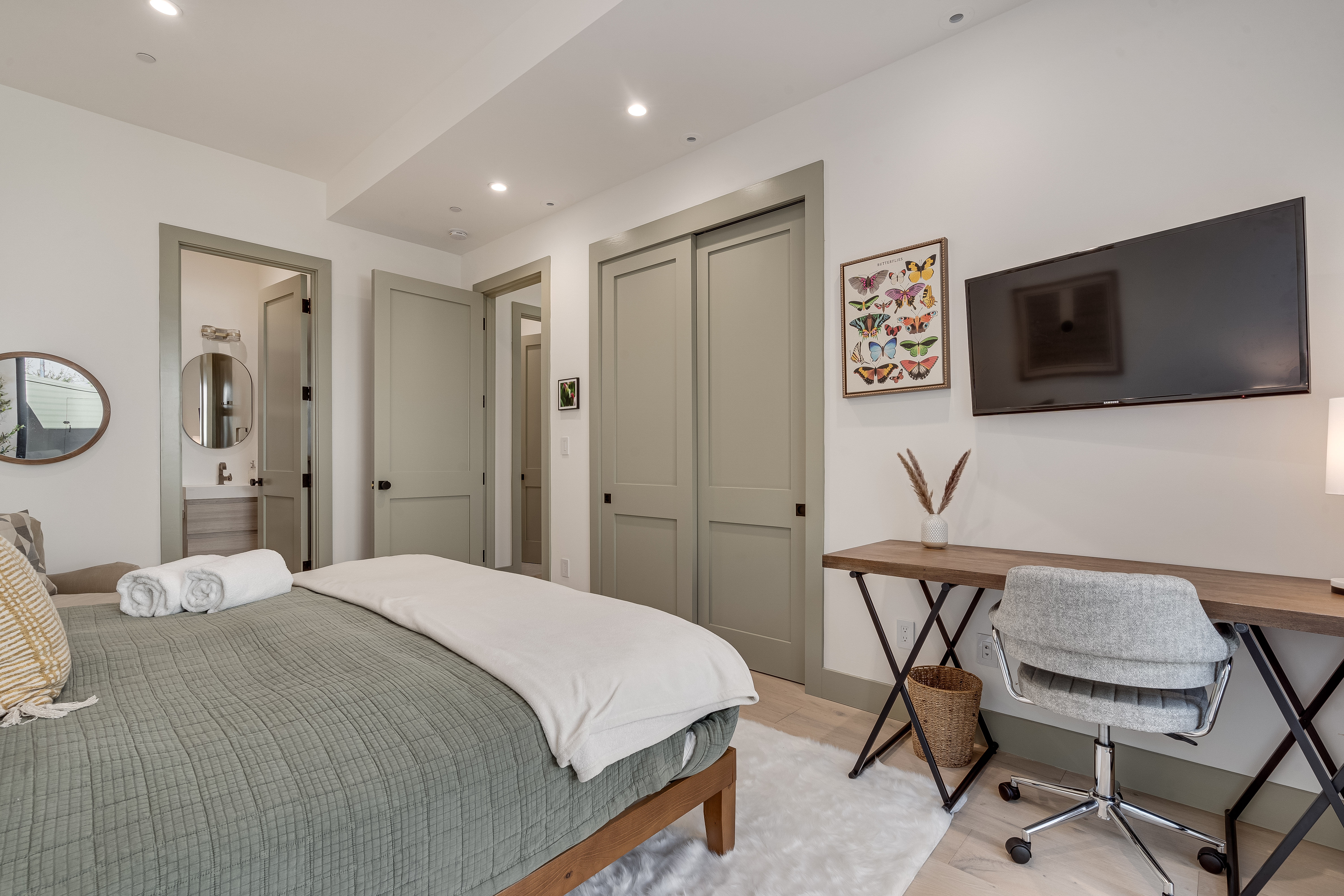 Contemporary bedroom interior with green quilted bed and rolled towels, linen throw and pillows, sliding closet doors, desk with swivel chair and wall-mounted TV, en-suite bathroom with round mirror, neutral tones and soft area rug.