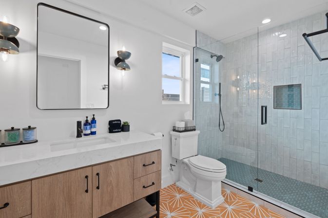 Bright modern bathroom with glass walk-in shower, blue mosaic shower floor and subway tile walls, white toilet, wood vanity with marble countertop and black-framed mirror, black-and-gold sconces and bold orange geometric floor tile.