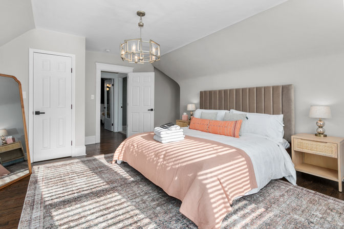 Sunlit modern master bedroom with a tall upholstered headboard, peach coverlet and orange lumbar pillow, crisp white sheets, wooden nightstands with lamps, geometric brass pendant, leaning full-length mirror, dark hardwood floors and patterned rug.