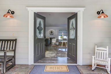 Welcoming front porch of a suburban home with open double black doors featuring decorative glass, a 'HELLO' doormat, copper wall sconces, wooden bench and white rocking chair, offering a view into a bright living room.