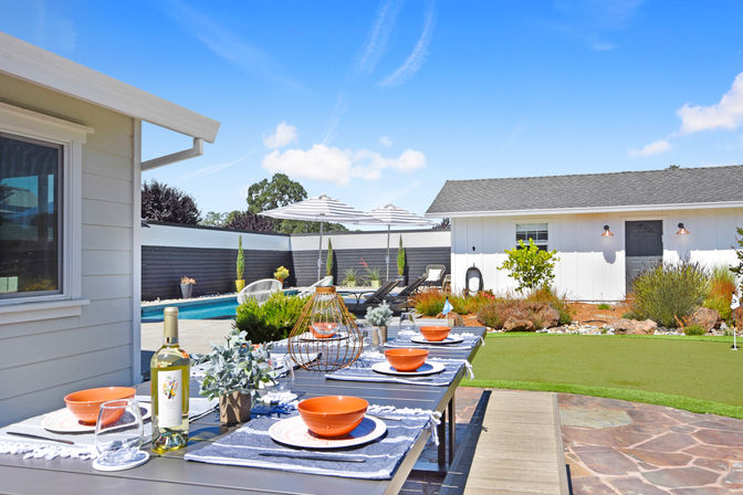 Sunny modern backyard patio with a long outdoor dining table set with orange bowls and a bottle of white wine, pool with striped umbrellas and lounge chairs, white pool house, putting green and landscaped garden.