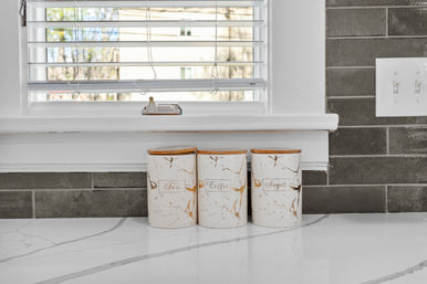 Three white ceramic canisters labeled Tea, Coffee, Sugar with bamboo lids on a white marble kitchen countertop beneath a window with blinds and gray subway tile backsplash.