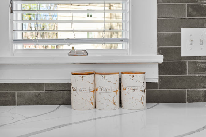 Three white ceramic canisters labeled Tea, Coffee, Sugar with bamboo lids on a white marble kitchen countertop beneath a window with blinds and gray subway tile backsplash.