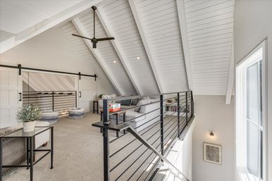 Bright modern loft living area with vaulted white shiplap ceiling, black metal railing and ceiling fan, sliding barn doors, gray sectional, pouf seats, and large window letting in natural light.